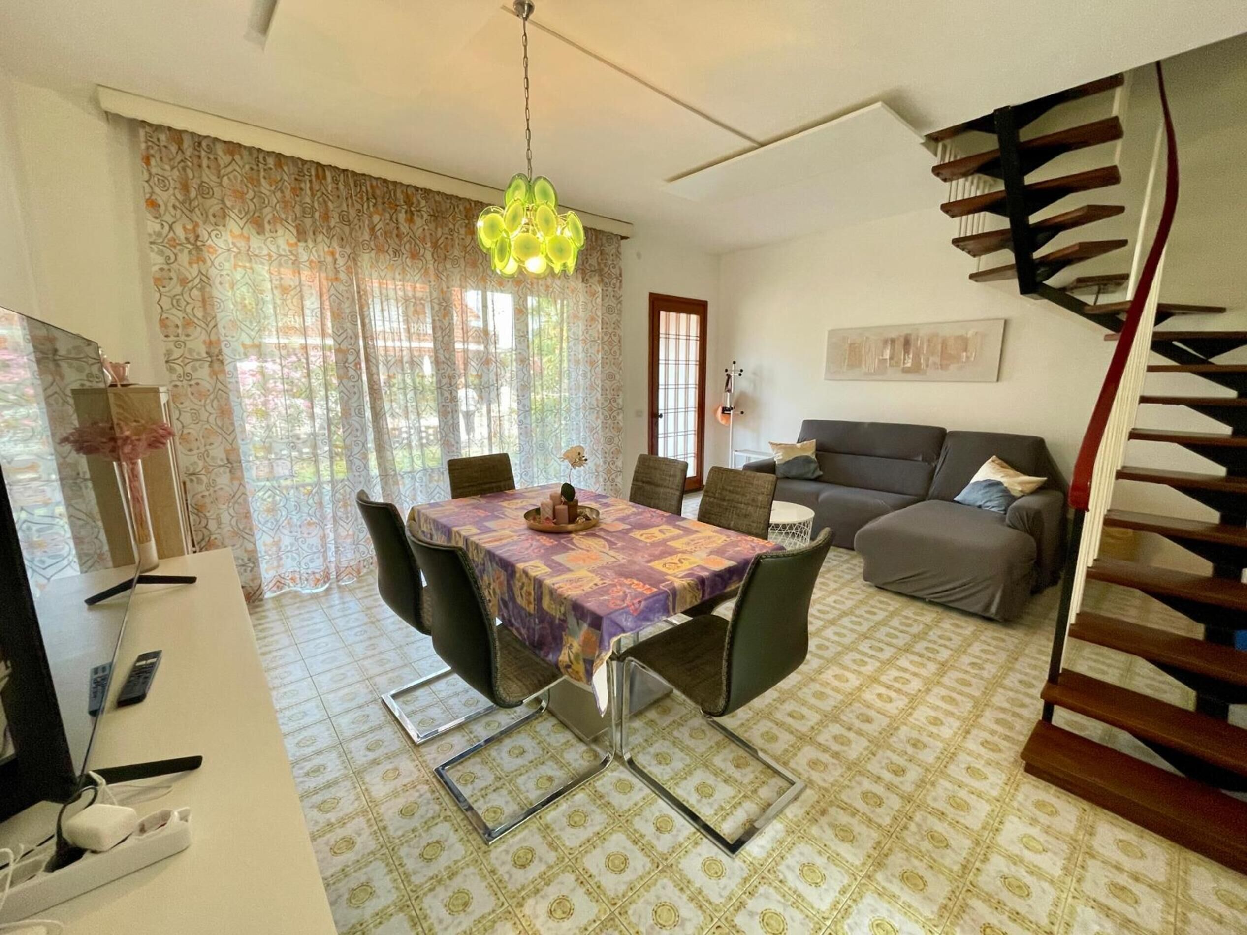 A dining area with a table set for four, a sofa, a staircase, and a large window with curtains.