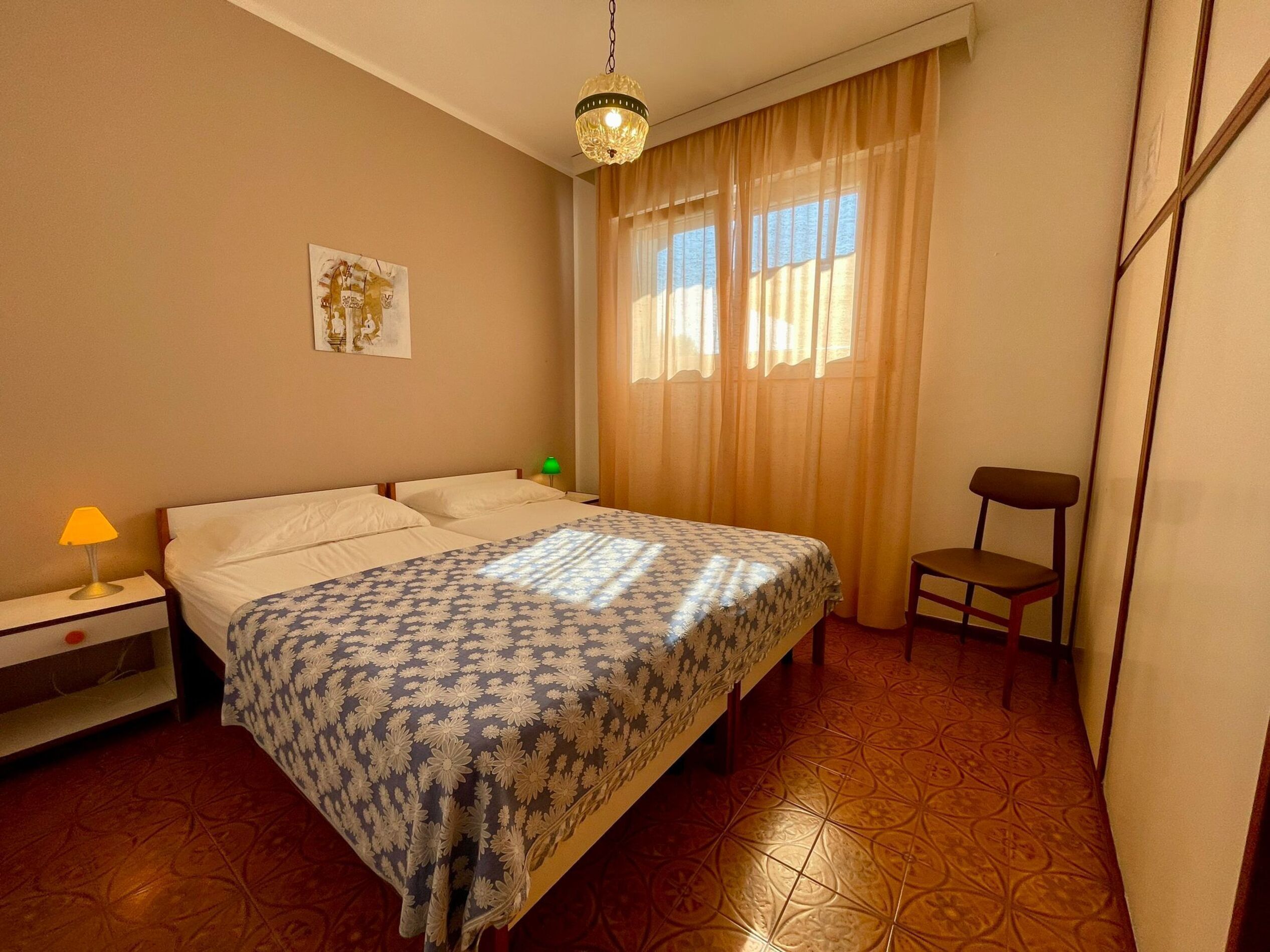 Un dormitorio con cama, mesita de noche, silla y ventana con cortinas.