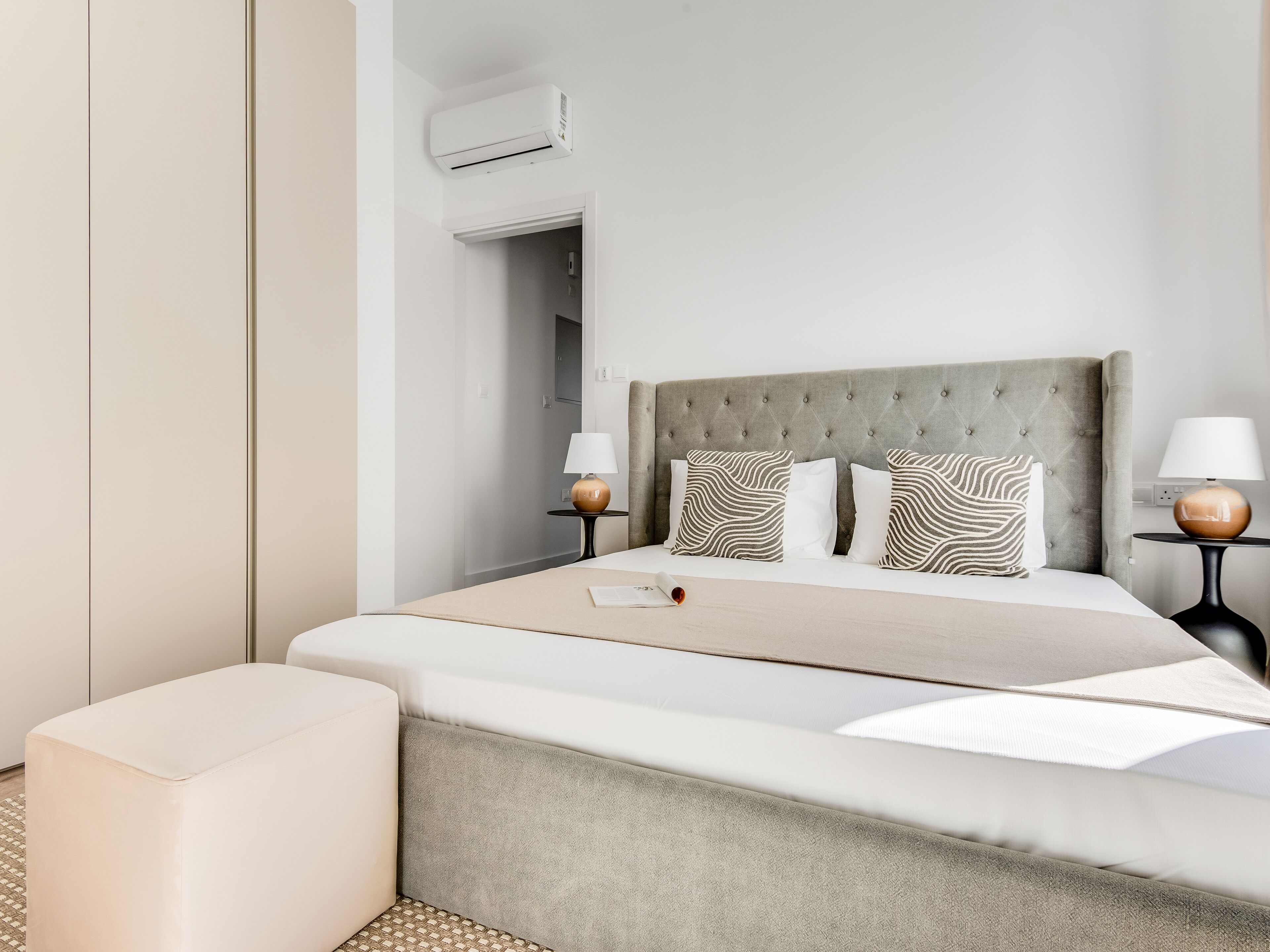 Una habitación de hotel moderna con una cama grande, dos mesitas de noche con lámparas, un cabecero tapizado en gris y una pequeña zona de estar.