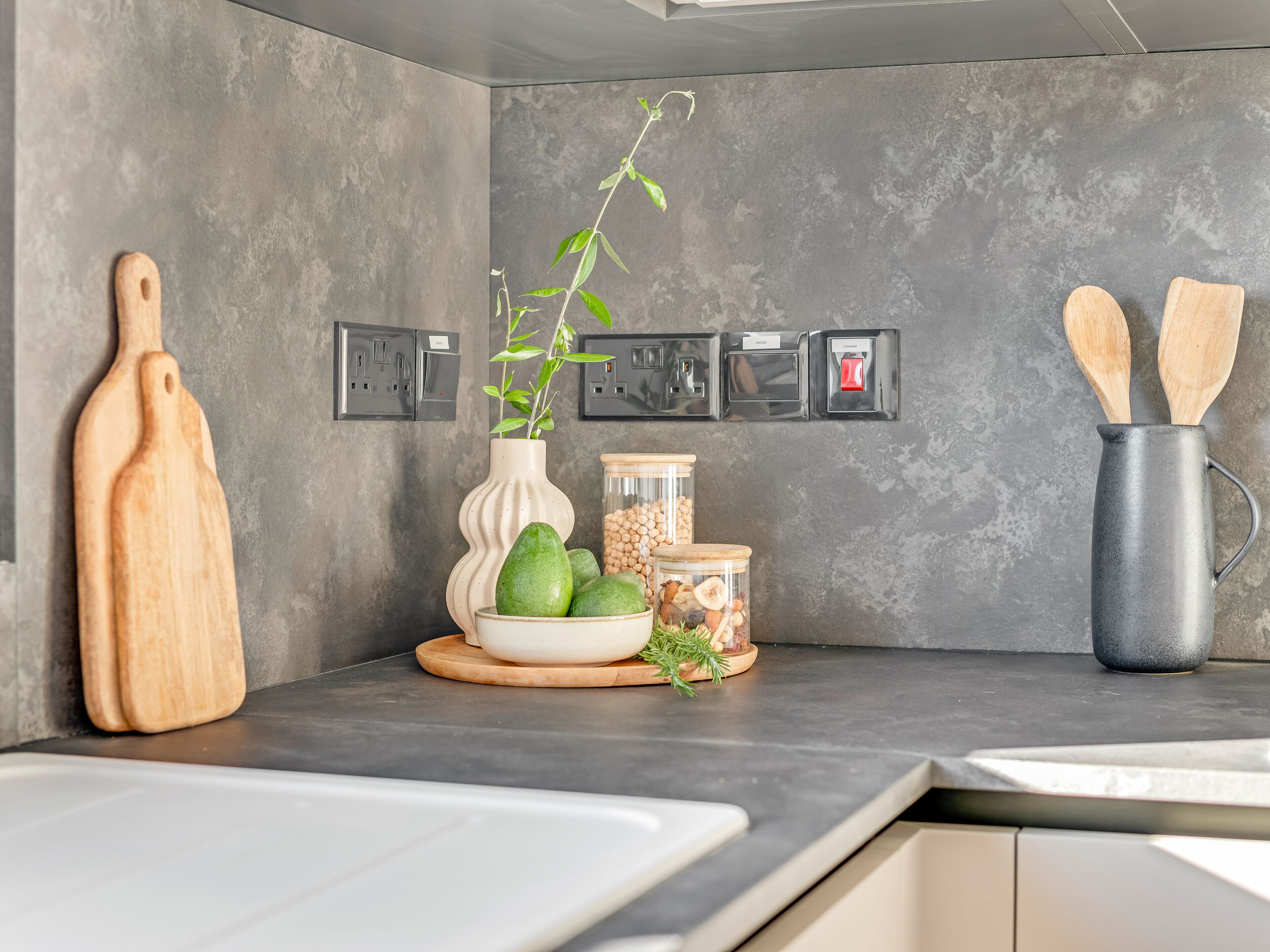Une cuisine avec un mur recouvert de carreaux gris, des planches à découper en bois, un vase avec des feuilles vertes, des bocaux remplis d’ingrédients et des cuillères en bois dans un pichet.