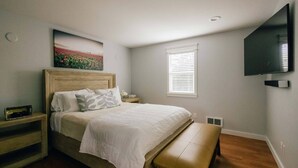 Un dormitorio con cama, banqueta, mesita de noche, televisor montado en la pared y ventana con persianas.
