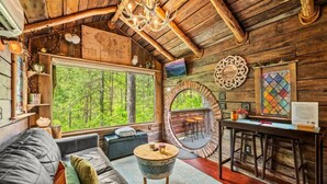 Interior rústico de una cabaña con un ventanal grande, un espejo redondo, techo de madera y una zona de estar acogedora.