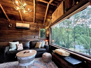 A cozy cabin interior with a leather sofa, a round wooden table, and a large window offering a forest view.