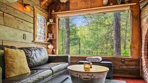 Interior de una cabaña con un sofá de cuero, una mesa redonda con una vela y un ventanal con vistas a un bosque.