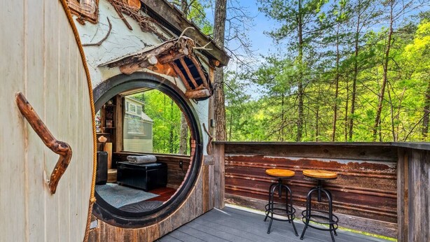 Una terraza de madera rústica con un ventanal circular, dos taburetes y vistas a un bosque.