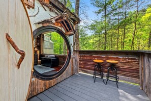 Une terrasse en bois rustique, dotée d’une fenêtre circulaire, de deux tabourets et offrant une vue sur une forêt.