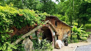 Un edificio de piedra rústico con un arco de entrada de madera, rodeado de abundante vegetación y un sendero de grava.