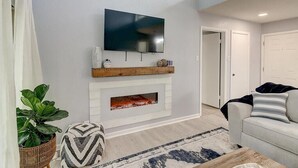 Ein Wohnzimmer mit Flachbildfernseher über einem Kamin, einer Couch, einem Couchtisch und einem Blumentopf.