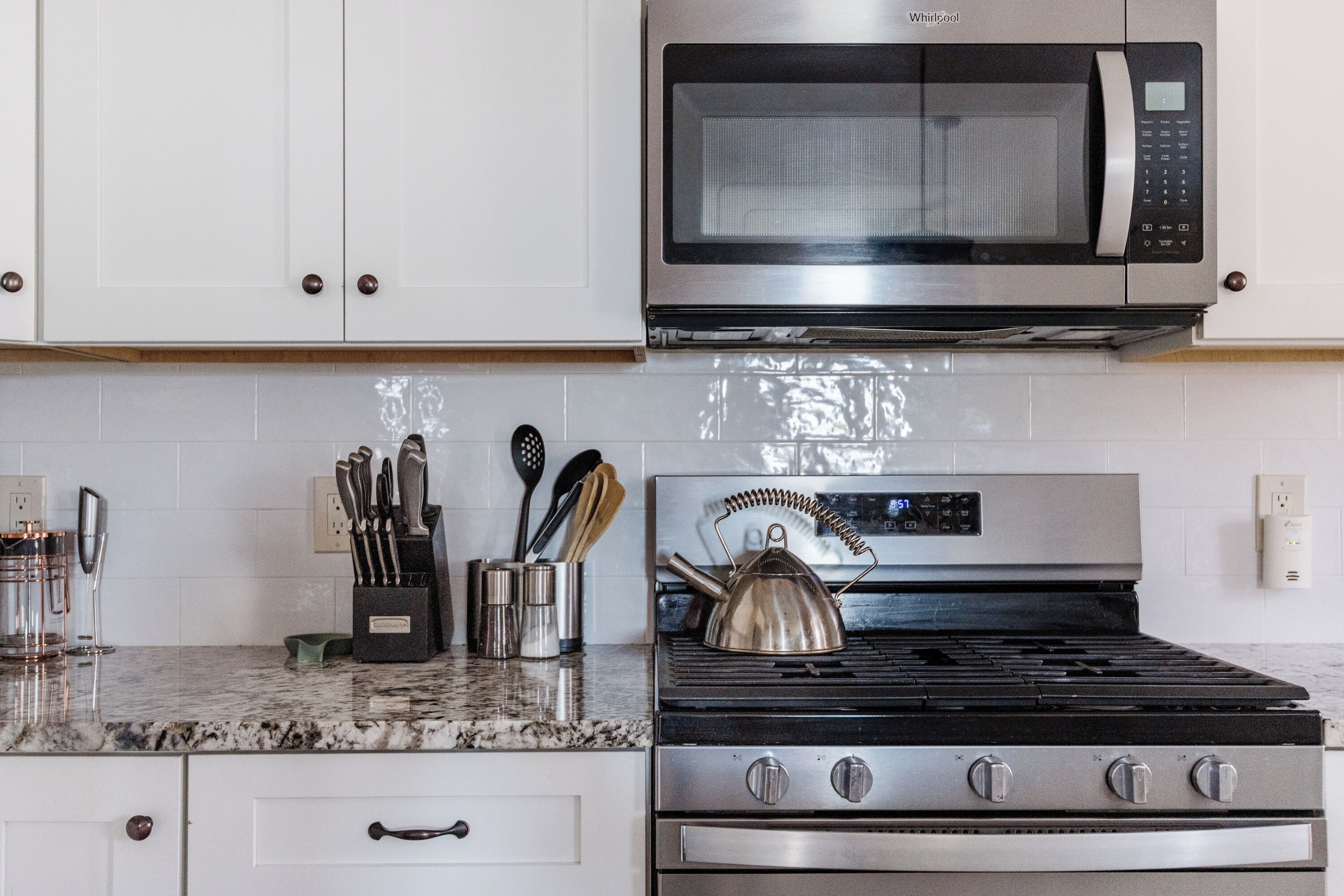 Una cocina con armarios blancos, un microondas de acero inoxidable, una estufa a gas y un hervidor de acero inoxidable.
