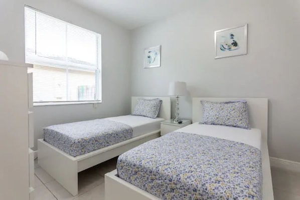 A bedroom with two single beds, a nightstand, a lamp, and two framed pictures on the wall. - Countryside Manor · Gateway to Keys & Everglades (Miami)