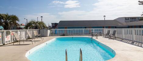A rectangular swimming pool with a no diving sign, surrounded by a white picket fence, and a building with a grey roof in the background.