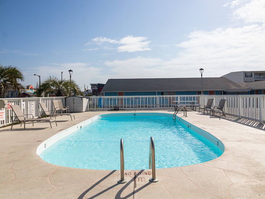 A rectangular swimming pool with a no diving sign, surrounded by a white picket fence, and a building with a grey roof in the background.