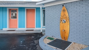 Um edifício com duas portas, uma laranja e outra azul, e uma prancha de surf encostada a uma parede de tijolos.