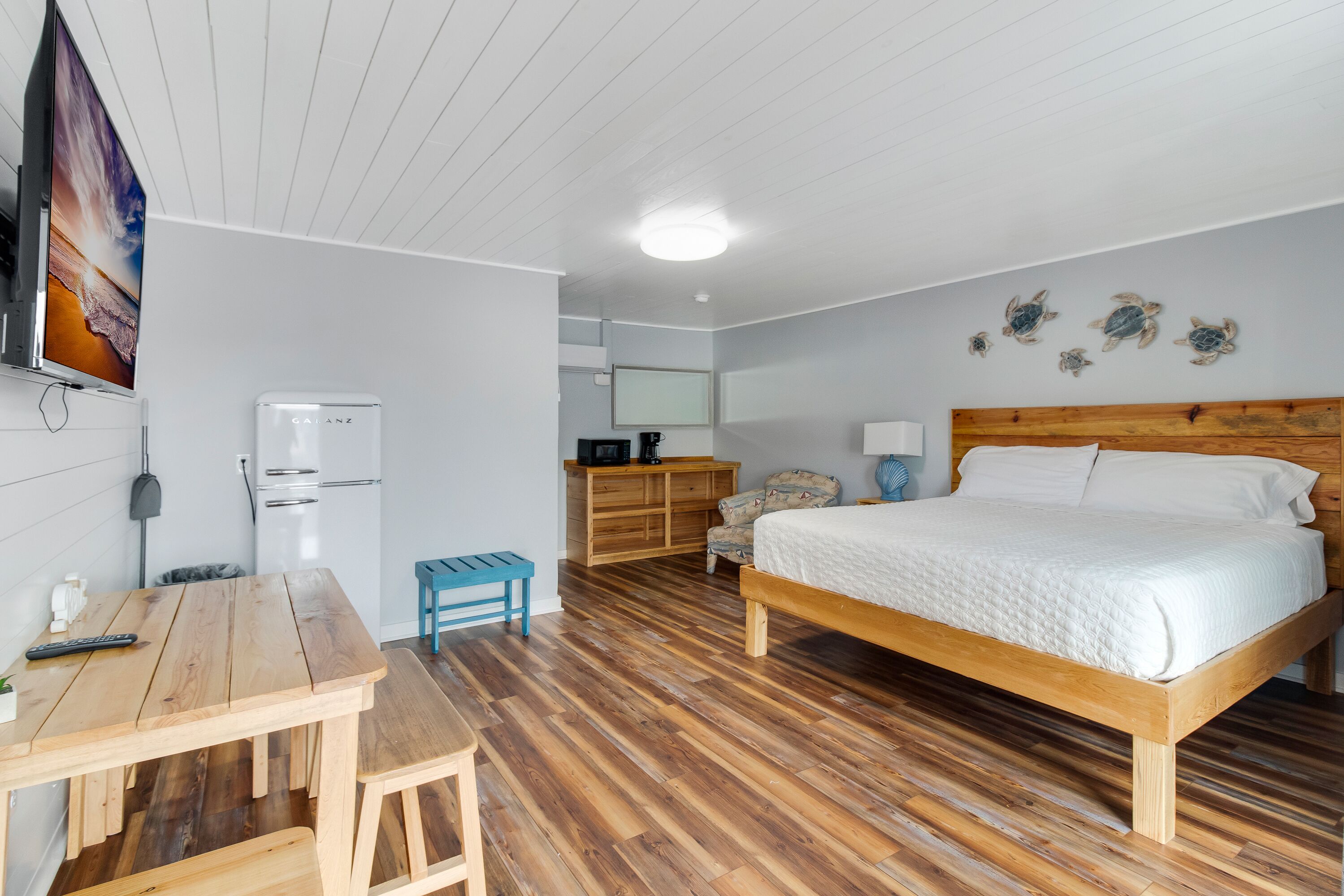 Un dormitorio con cama de madera, un escritorio de madera, una mesita pequeña, una silla, un refrigerador, una cafetera y un televisor montado en la pared.