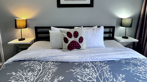 A neatly made bed with white bedding and a tree branch pattern, two bedside lamps, a framed picture of mountains, and a white pillow with a paw print design.