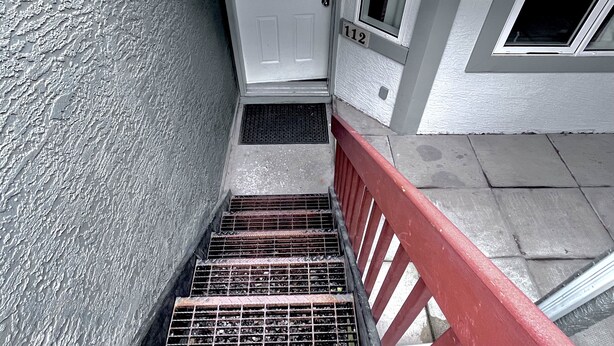 A stairway with a metal grate at the bottom, a red handrail, and a white door with the number 112. - Two-Bedroom Apartment 131 - Private Entrance - Self Check In (BL-RES-12552) (Canmore)