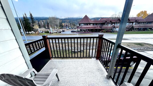 Un balcon avec deux chaises en bois, une vue sur un parking et, au loin, un immeuble avec un toit rouge.