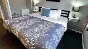 Un lit bien fait, recouvert d’une couvre-lit à motifs, deux tables de chevet avec des lampes, un tableau encadré au mur et une armoire avec un miroir.