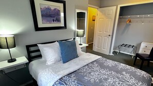 Un lit bien fait, recouvert d’une couvre-lit à motifs floraux blancs et bleus, deux lampes de chevet, un tableau encadré au mur et un dressing avec un miroir.
