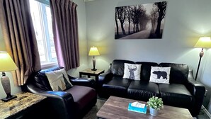 Ett vardagsrum med en svart lädersoffa, ett träbord och ett litet sidobord. Det finns ett fönster med gardiner, en lampa på sidobordet och en tavla på väggen.