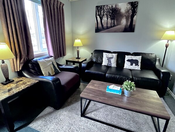 A living room with a black leather sofa, a wooden coffee table, and a small side table. There is a window with curtains, a lamp on the side table, and a painting on the wall. - Three-Bedroom #116 - Private Entrance - Self Check In-Railside. BL-RES-12551. (Canmore)
