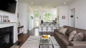 A modern living room with a fireplace, a glass coffee table with fruit, a brown sofa, and a kitchen area with bar stools.