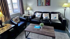 Ein Wohnzimmer mit einer schwarzen Ledersofa, zwei Sesseln, einem Couchtisch und einem kleinen Beistelltisch mit einer Lampe. Ein Fenster mit Vorhängen, ein Gemälde an der Wand und eine Topfpflanze auf dem Couchtisch sind ebenfalls zu sehen.