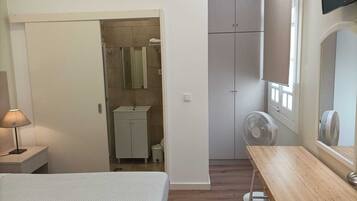 A bedroom with a bed, a bedside table, a wooden desk, a mirror, a wardrobe, a bathroom with a sink, and a fan.