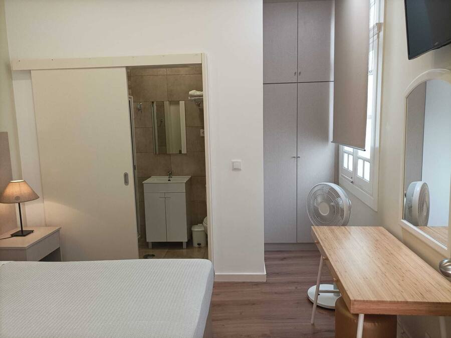 A bedroom with a bed, a bedside table, a wooden desk, a mirror, a wardrobe, a bathroom with a sink, and a fan.