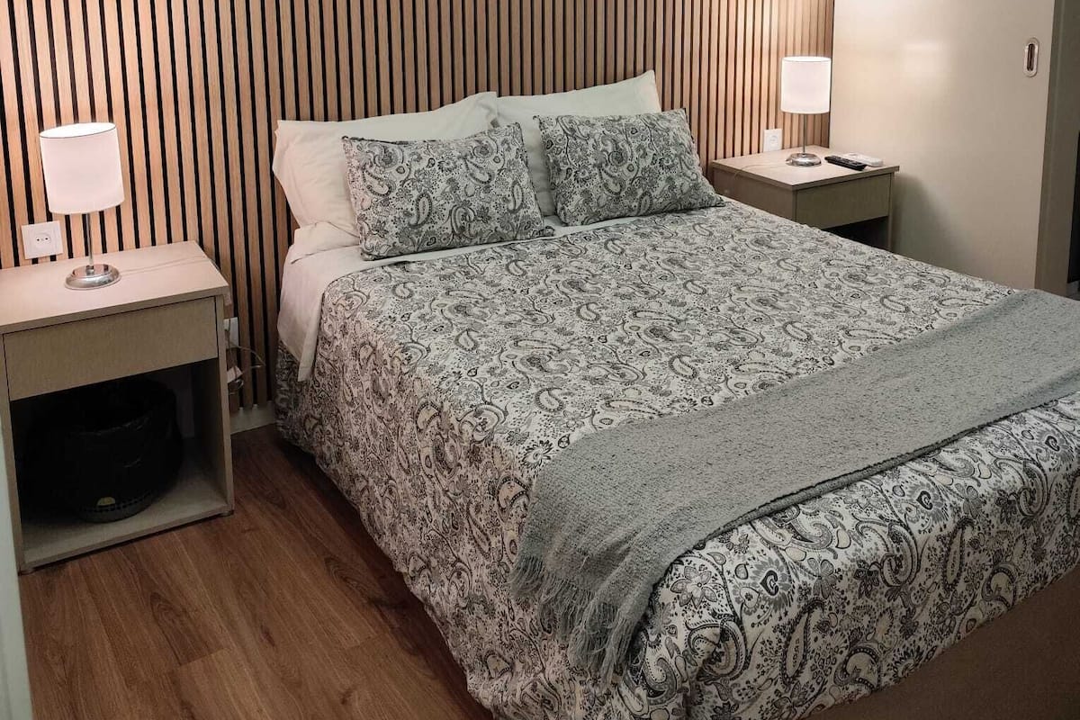 A neatly arranged bedroom with a bed, two bedside tables, a wall-mounted air conditioner, and a decorative wall piece.