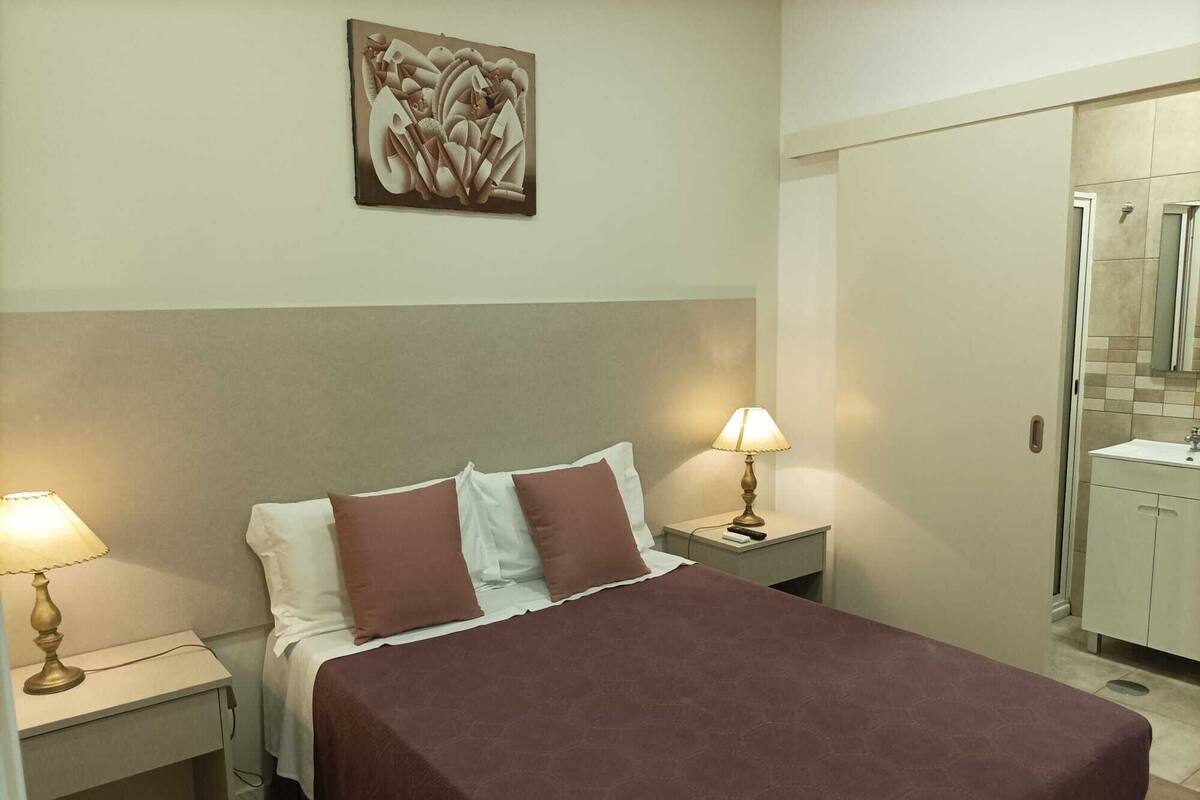 A hotel room with a bed, two bedside lamps, a nightstand, a wall-mounted artwork, and a bathroom visible through an open door.