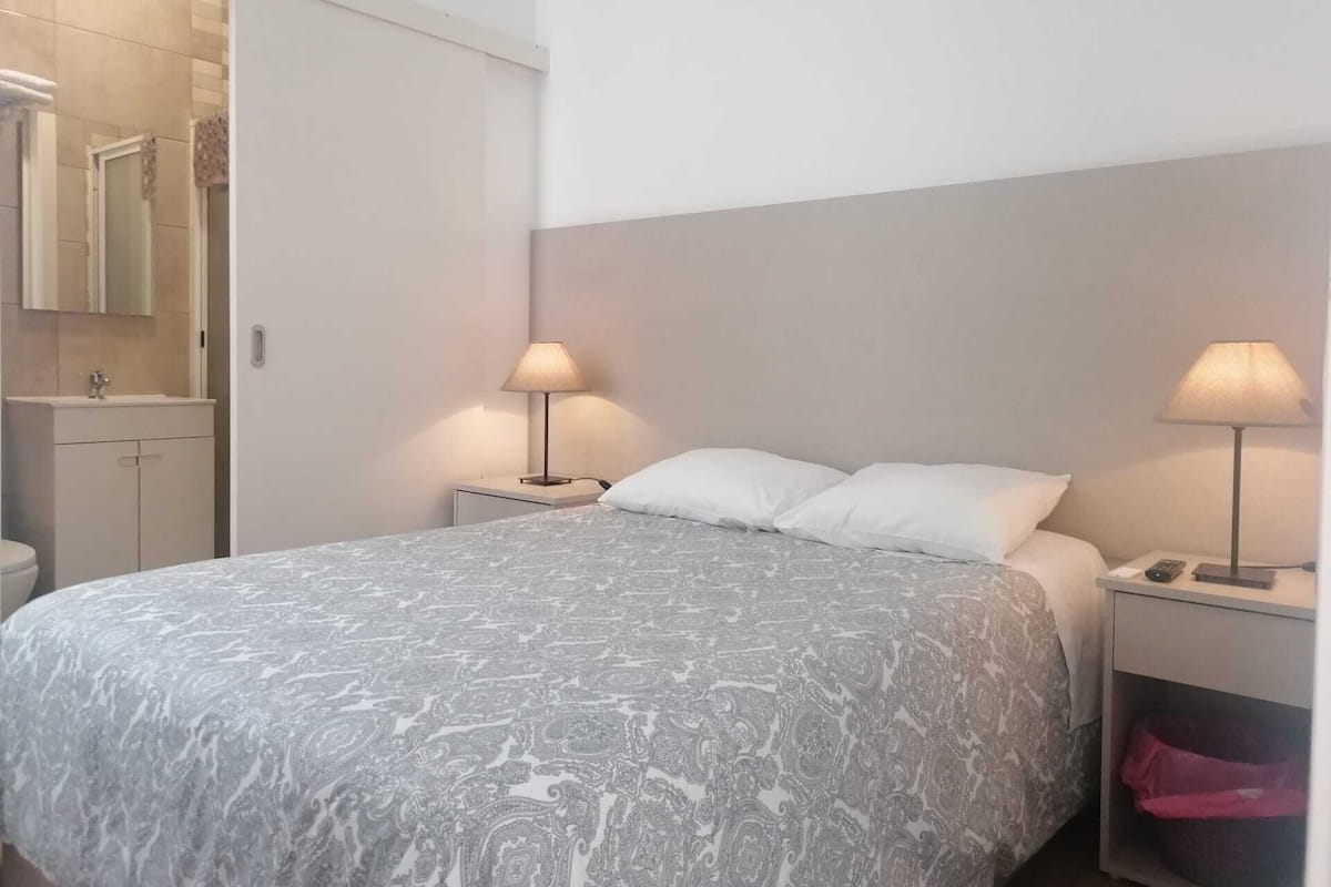 A bedroom with a bed, two bedside lamps, a nightstand, a small drawer, and a door leading to a bathroom.