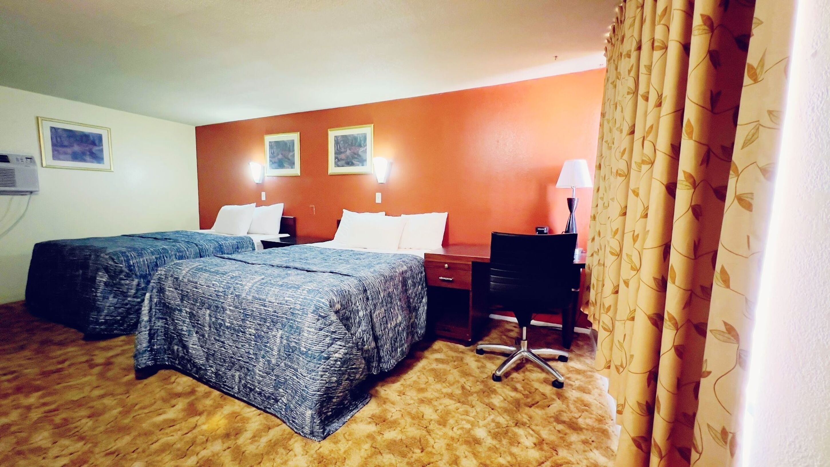 A hotel room with two beds, a desk, a chair, and a lamp.