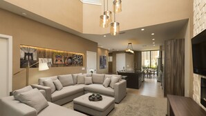 Una sala de estar moderna con un sofá seccional, una mesa de centro y un televisor de pantalla plana. Se observa un área de cocina al fondo con una mesa de comedor y sillas.