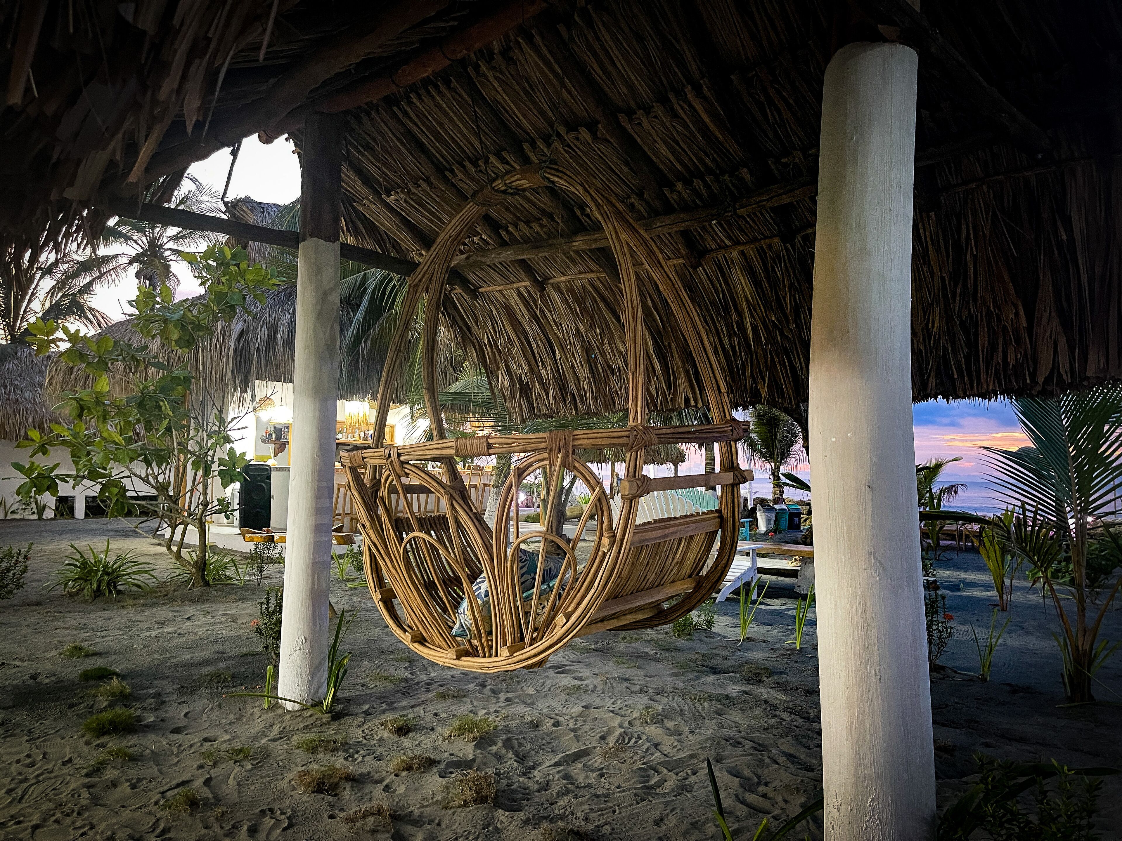 A beachside structure with a thatched roof, a hanging wicker swing, and a sandy ground with sparse vegetation.
