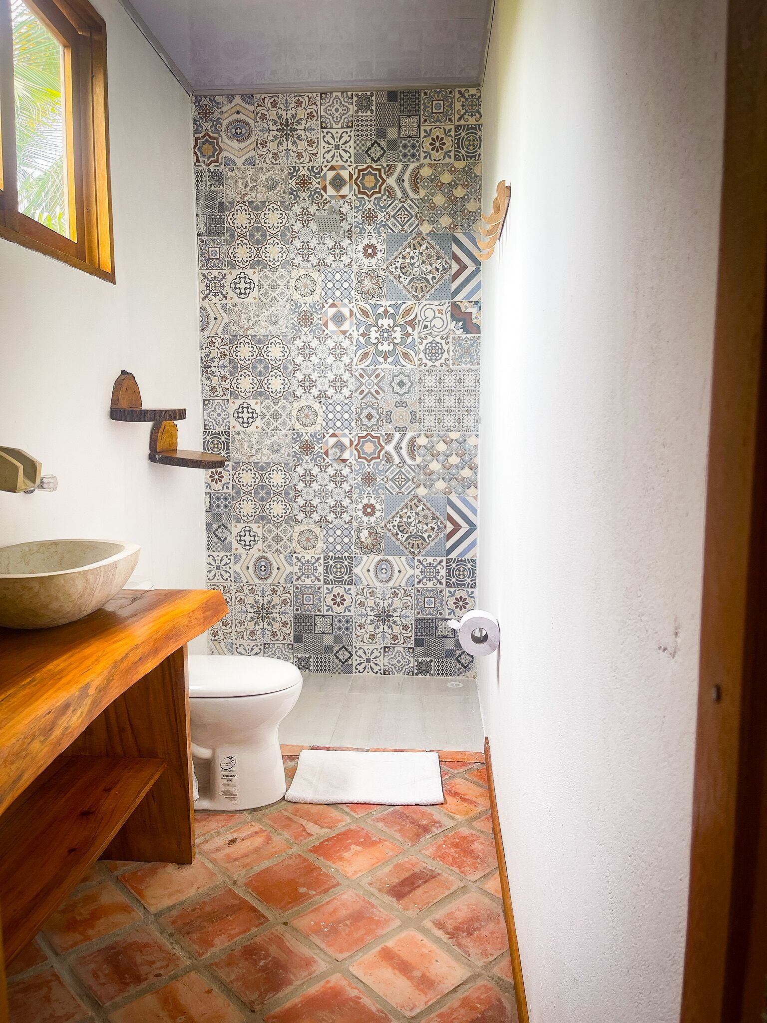 A bathroom with a tiled shower wall, a wooden vanity, a white toilet, and a red-tiled floor.