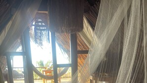 A room with a bed covered in white netting, wooden flooring, and a view of an outdoor area with a thatched roof and wooden furniture. - CASA ORIGEN BOUTIQUE (San Bernardo del Viento)