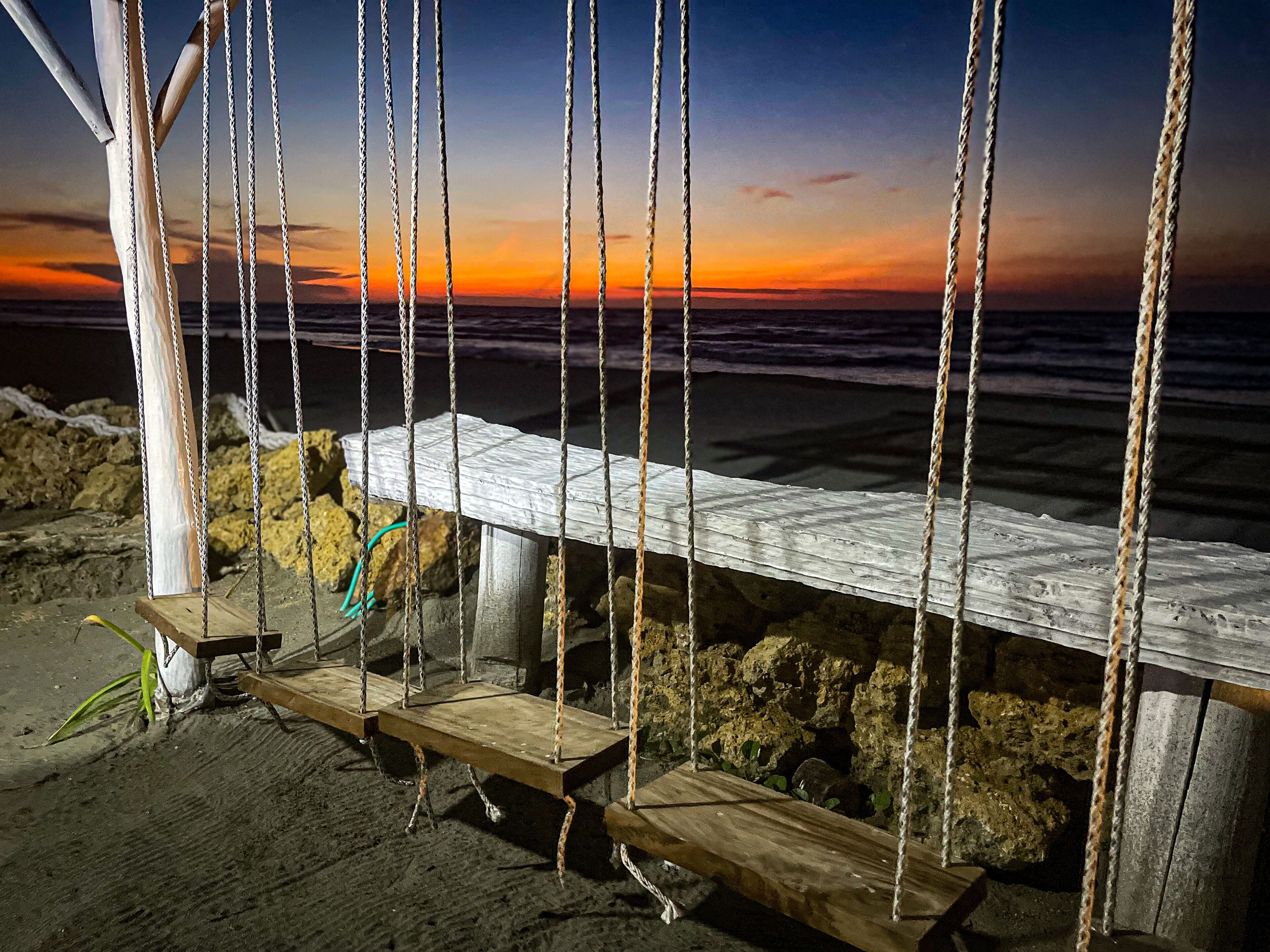 Un conjunto de columpios de madera con cuerdas, ubicado en una playa arenosa cerca de un afloramiento rocoso.