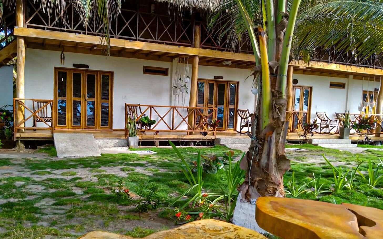 Un edificio de dos plantas con techo de paja, balcones de madera y un jardín con plantas tropicales.