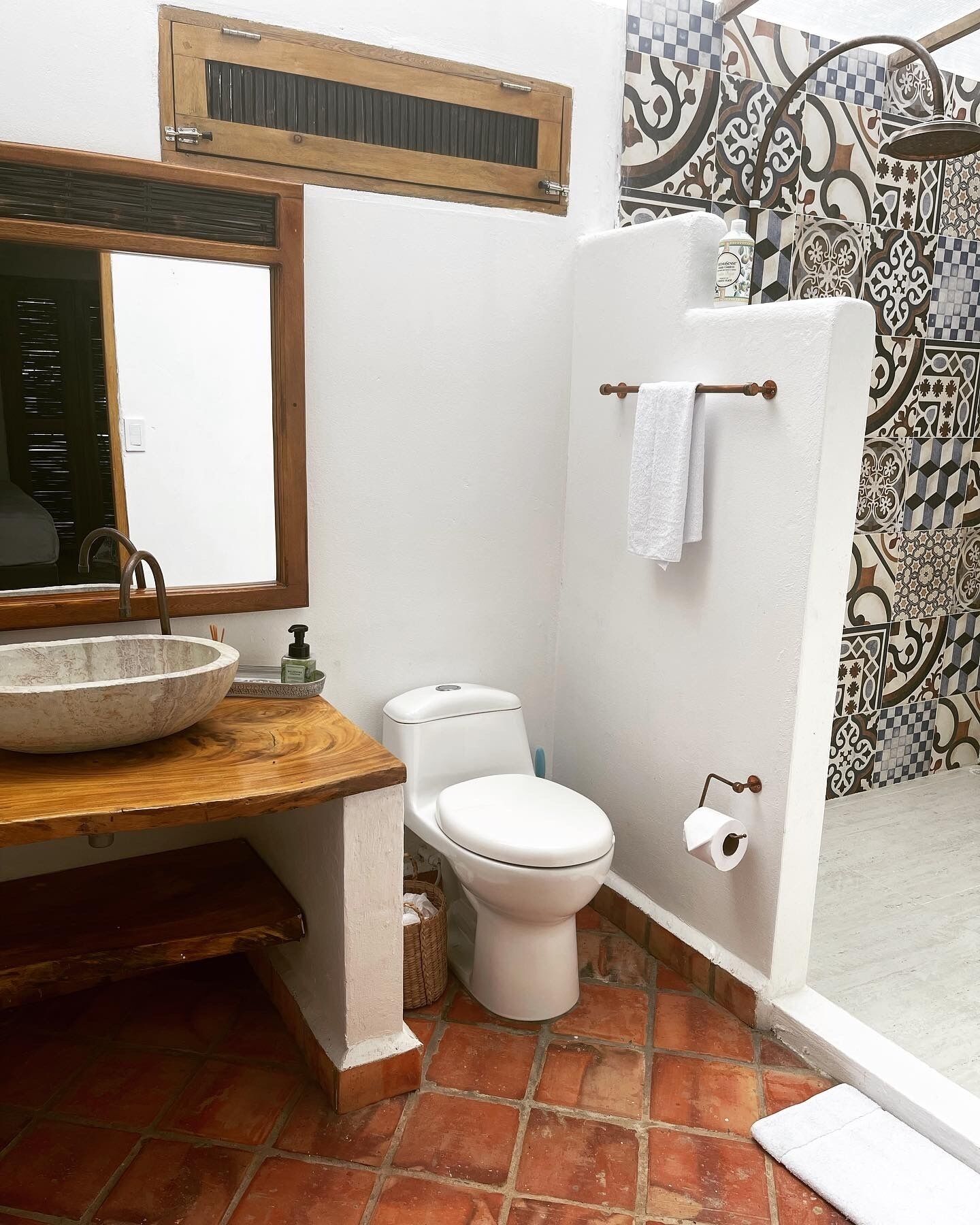 Une salle de bain avec un meuble-lavabo en bois, des toilettes blanches, une douche avec un mur carrelé et une fenêtre avec un cadre en bois.