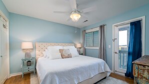 A bedroom with a bed, two bedside tables with lamps, a ceiling fan, a window with curtains, and a door with a view.