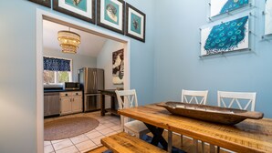 A dining area with a wooden table, benches, and a chandelier. There is a kitchen visible in the background with cabinets and a microwave.