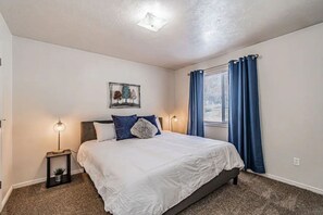 Un dormitorio con una cama, dos mesitas de noche, una lámpara de noche, una ventana con cortinas y una lámpara en el techo.