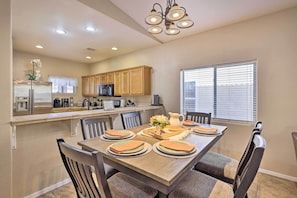 Un comedor con una mesa de madera dispuesta para cuatro personas, rodeada de sillas. Se observa una cocina al fondo con armarios de madera y un refrigerador.