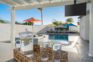 A poolside setting with a clear glass pitcher and glasses on a woven tray, a pool, and umbrellas labeled "COACHELL" in the background. - Luxurious desert home w/ pool & jacuzzi! (Coachella)