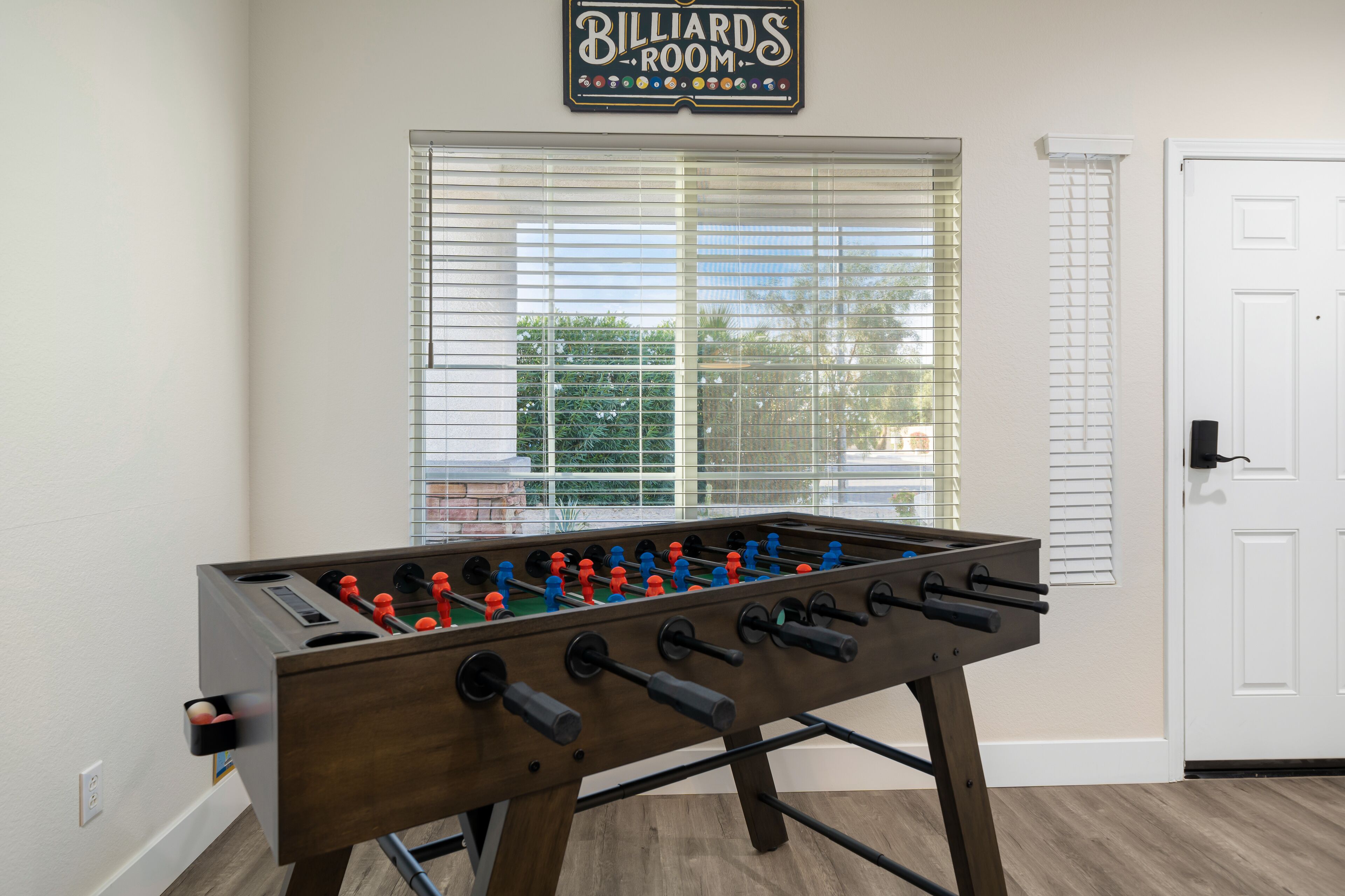 A billiards room with a foosball table, a window with blinds, and a white door.