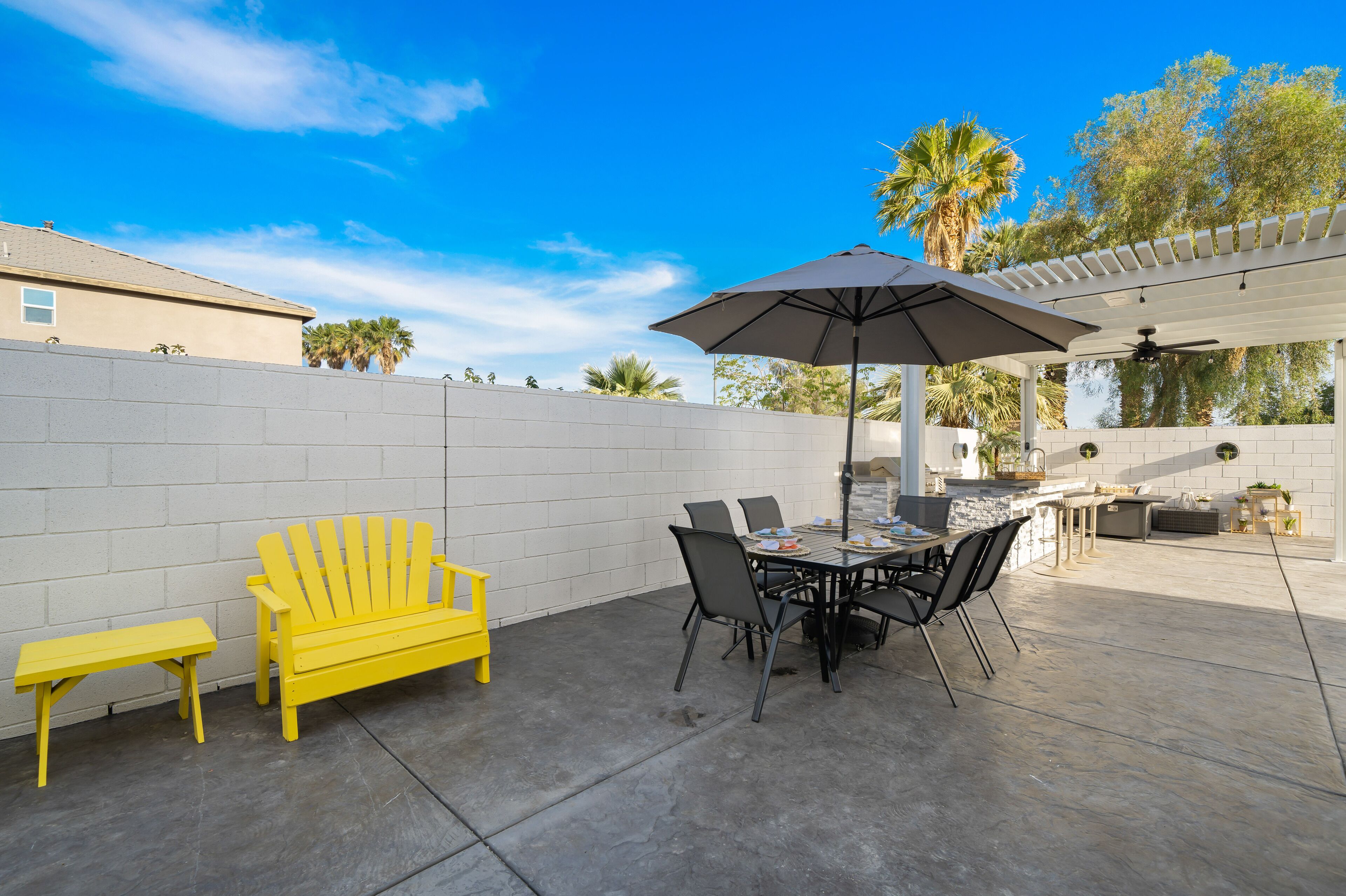 An outdoor patio area with a table and chairs, a yellow Adirondack chair, and a pergola with an umbrella.