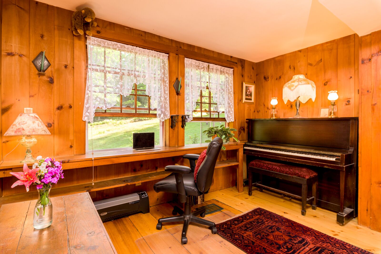 A cozy room with wooden walls, a piano, a desk with a laptop, a chair, and a window with lace curtains.