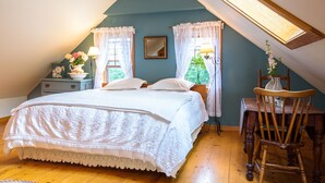 Une chambre avec un plafond en pente, un lit recouvert de draps blancs, deux fenêtres ornées de rideaux légers, une table de chevet avec une lampe et une chaise sur laquelle est posé un vase de fleurs.