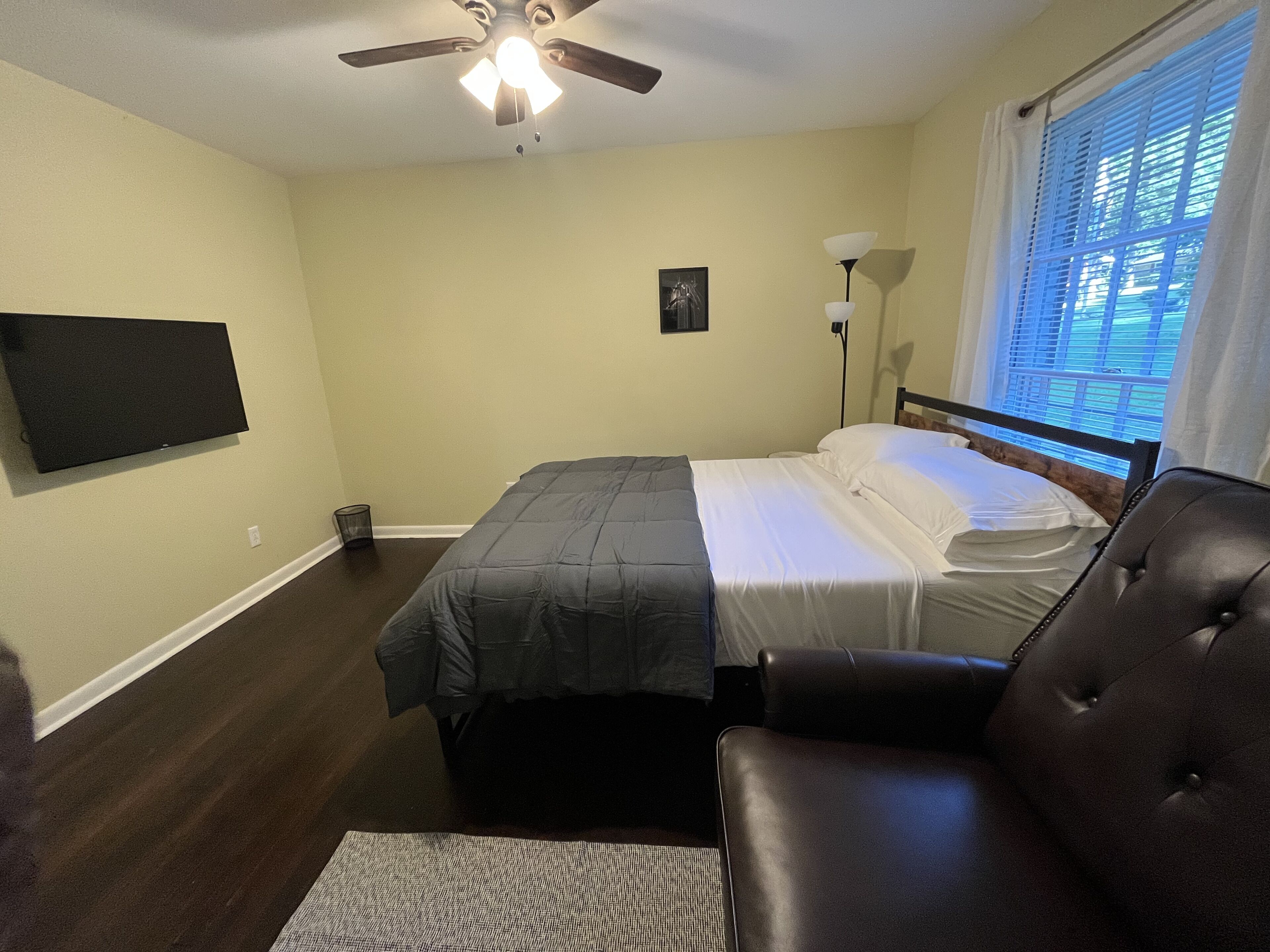A bedroom with a bed, a chair, a ceiling fan, a TV, and a window with blinds.